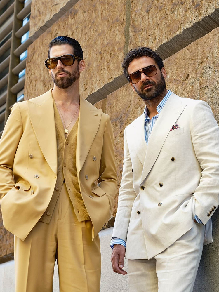 Two men in tailored double-breasted suits, one in sand with a matching cardigan and one in cream linen with a striped shirt