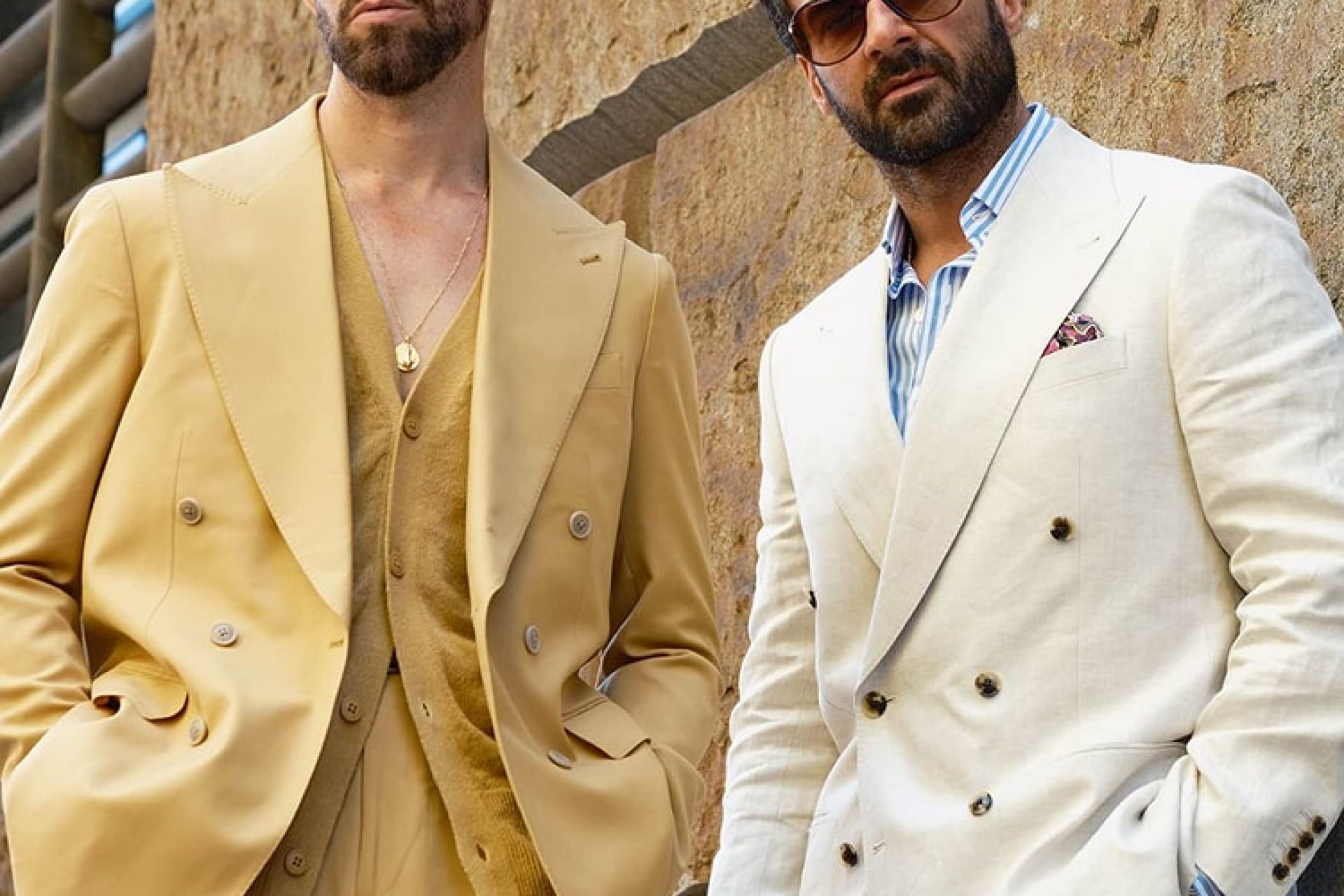 Two men in tailored double-breasted suits, one in sand with a matching cardigan and one in cream linen with a striped shirt