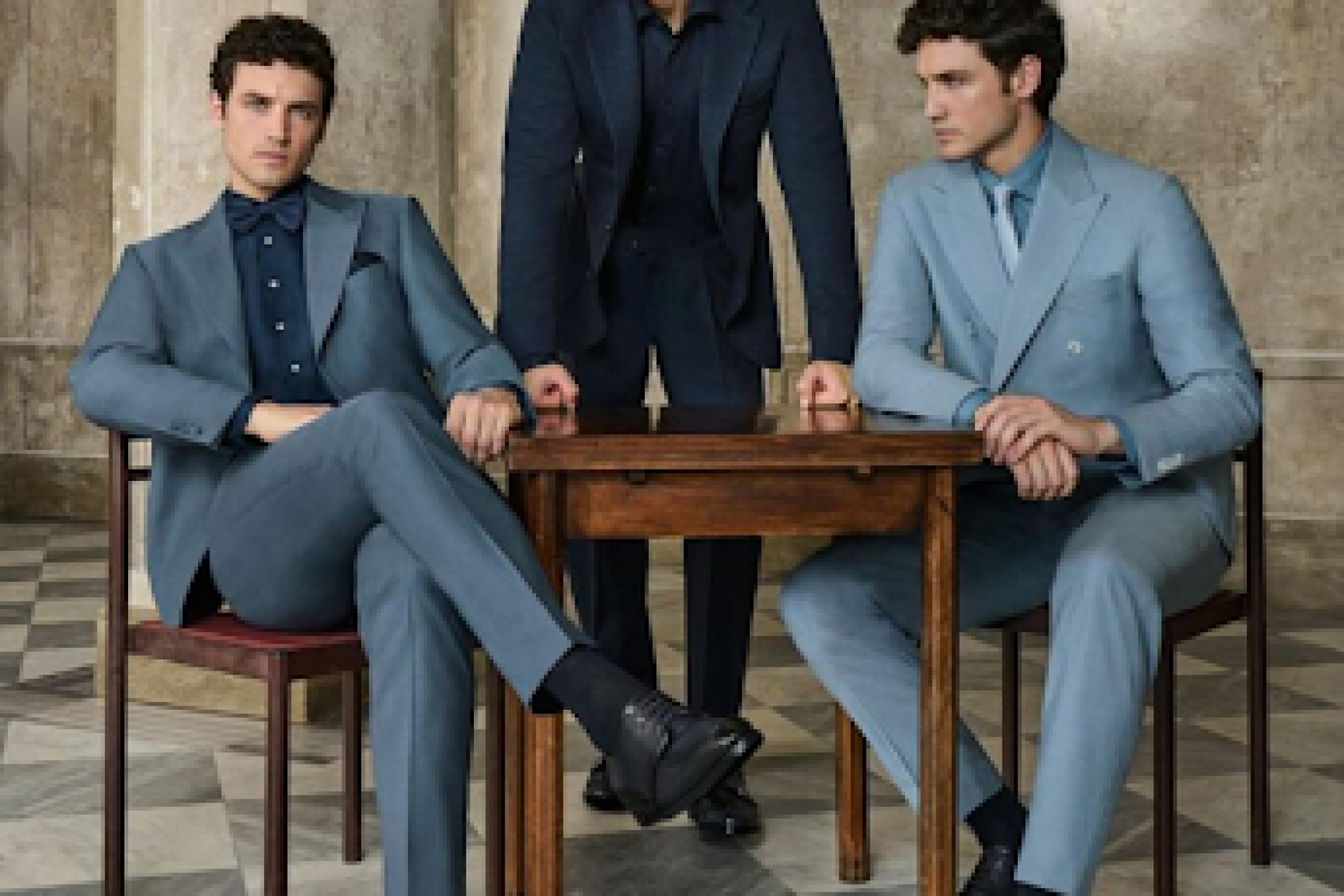Three men wearing well-kept tailored suits in navy, slate, and light blue around a wooden table